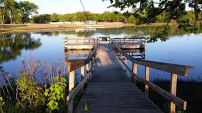 A wooden boardwalk extends out over a calm, reflective lake, leading to a small fishing pier with railings and benches. The water mirrors the surrounding green trees and blue sky, creating a peaceful, scenic view. Tall grasses and foliage frame the edge of the shoreline, adding to the natural, tranquil setting.